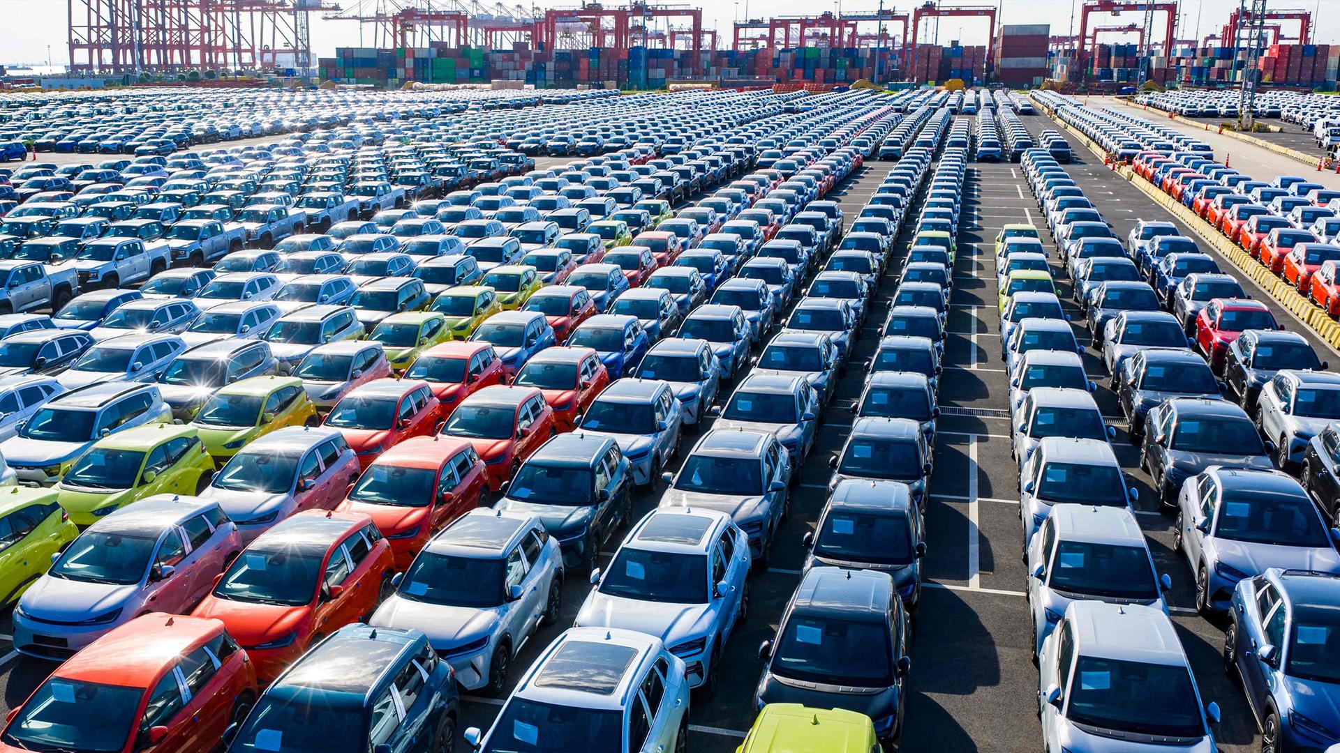 Reihen neuer Autos auf einem großen Hafengelände mit Containern und Kränen im Hintergrund.
