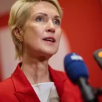 Eine blonde Frau spricht bei einer Pressekonferenz vor mehreren Mikrofonen.