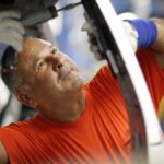 Ein Arbeiter in orangefarbenem T-Shirt montiert in einer Werkstatt ein Bauteil an einem Fahrzeug.