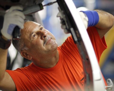 Ein Arbeiter in orangefarbenem T-Shirt montiert in einer Werkstatt ein Bauteil an einem Fahrzeug.