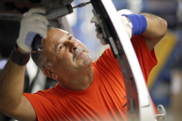 Ein Arbeiter in orangefarbenem T-Shirt montiert in einer Werkstatt ein Bauteil an einem Fahrzeug.