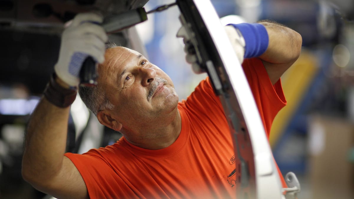 Ein Arbeiter in orangefarbenem T-Shirt montiert in einer Werkstatt ein Bauteil an einem Fahrzeug.