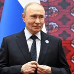 Wladimir Putin steht in einem dunklen Anzug vor russischen und anderen Flaggen.