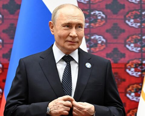 Wladimir Putin steht in einem dunklen Anzug vor russischen und anderen Flaggen.