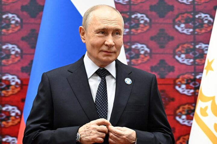 Wladimir Putin steht in einem dunklen Anzug vor russischen und anderen Flaggen.