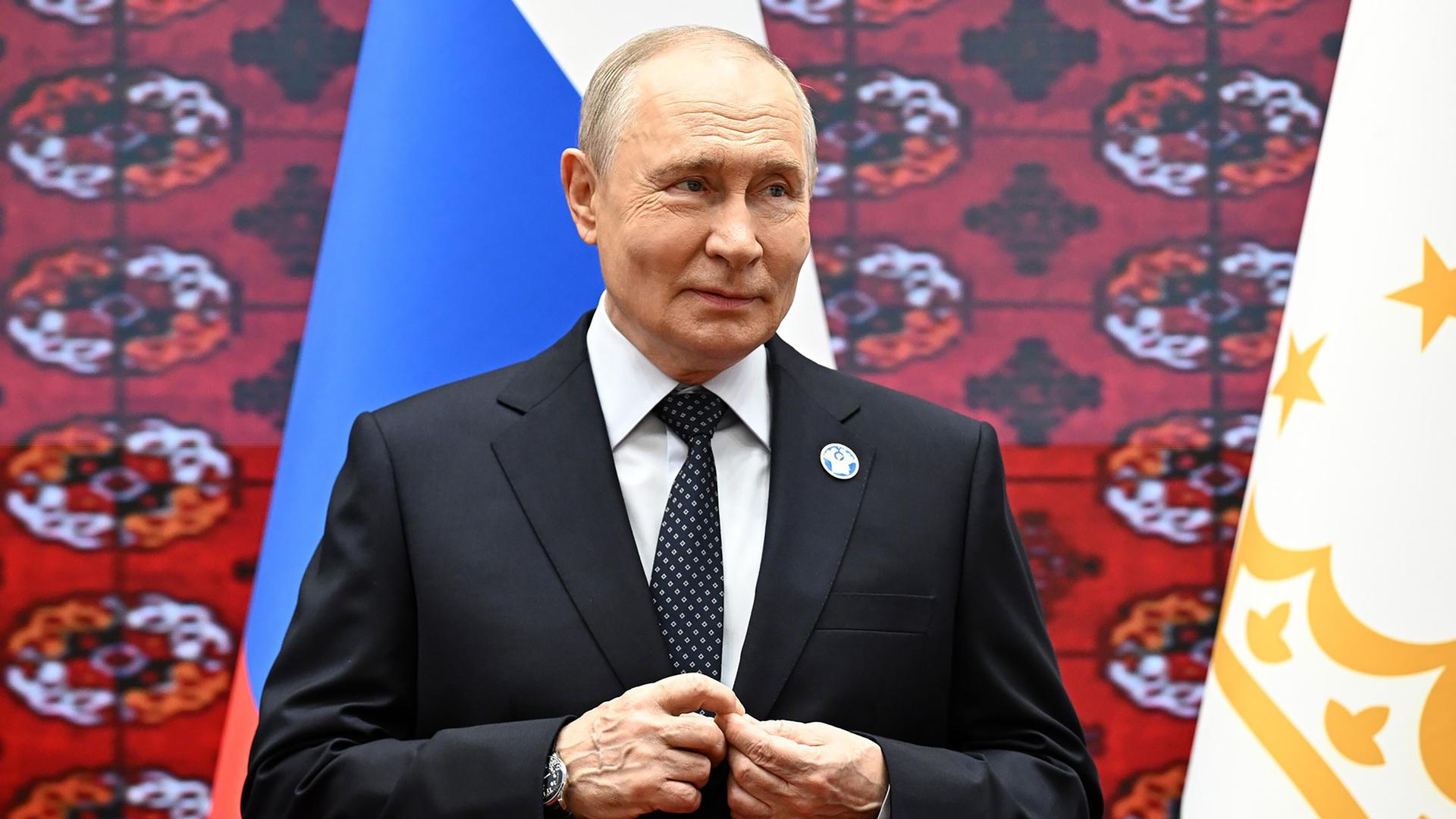 Trump-Diesel-Deal stärkt Putin laut Bericht politisch und wirtschaftlich 1 Wladimir Putin steht in einem dunklen Anzug vor russischen und anderen Flaggen.