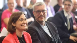 Zwei Politiker sitzen bei einer Veranstaltung und lächeln in den Raum.