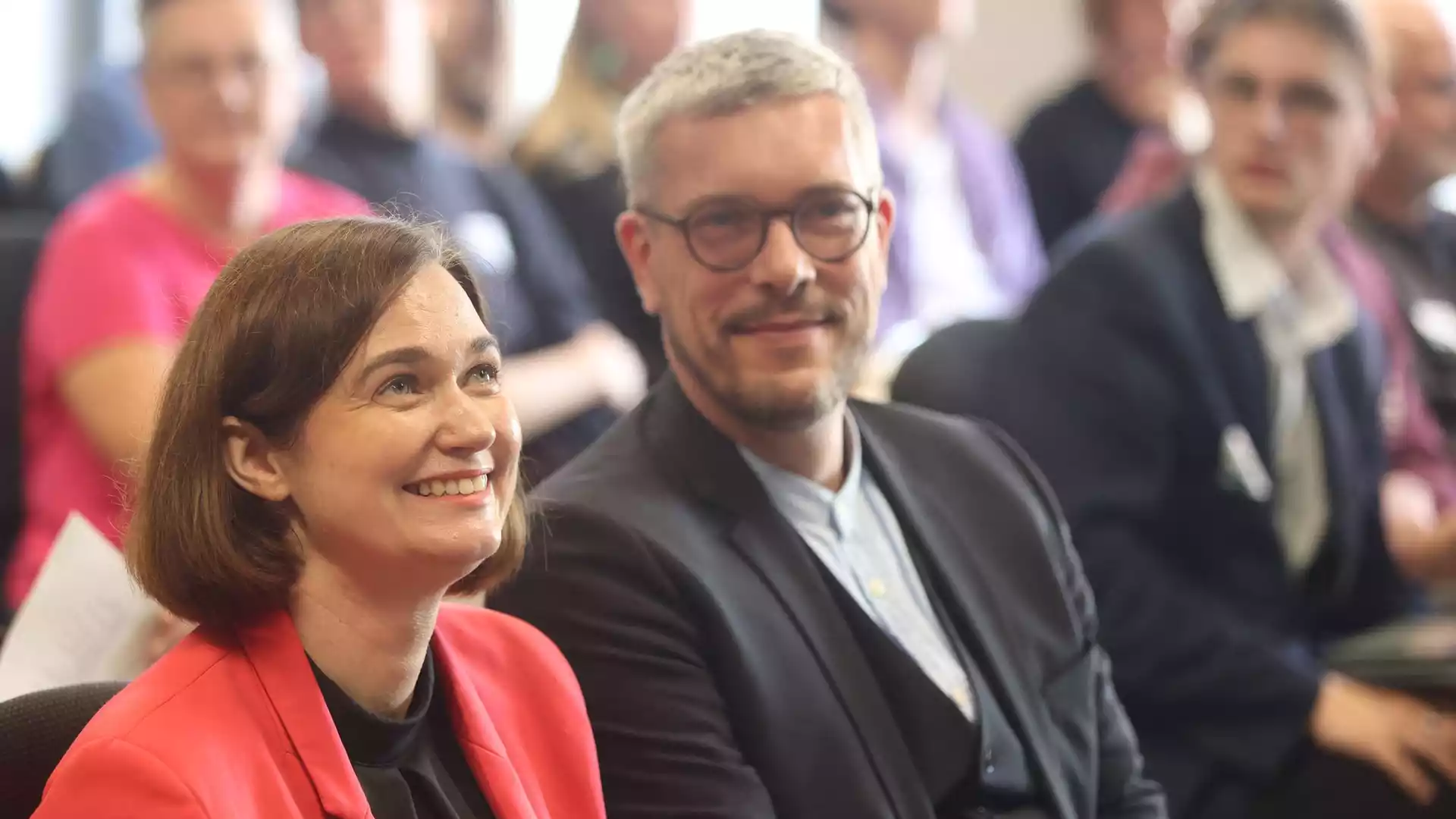 Zwei Politiker sitzen bei einer Veranstaltung und lächeln in den Raum.