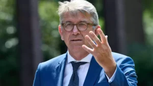 Ein Mann im blauen Anzug und mit Brille spricht im Freien und hebt die Hand.