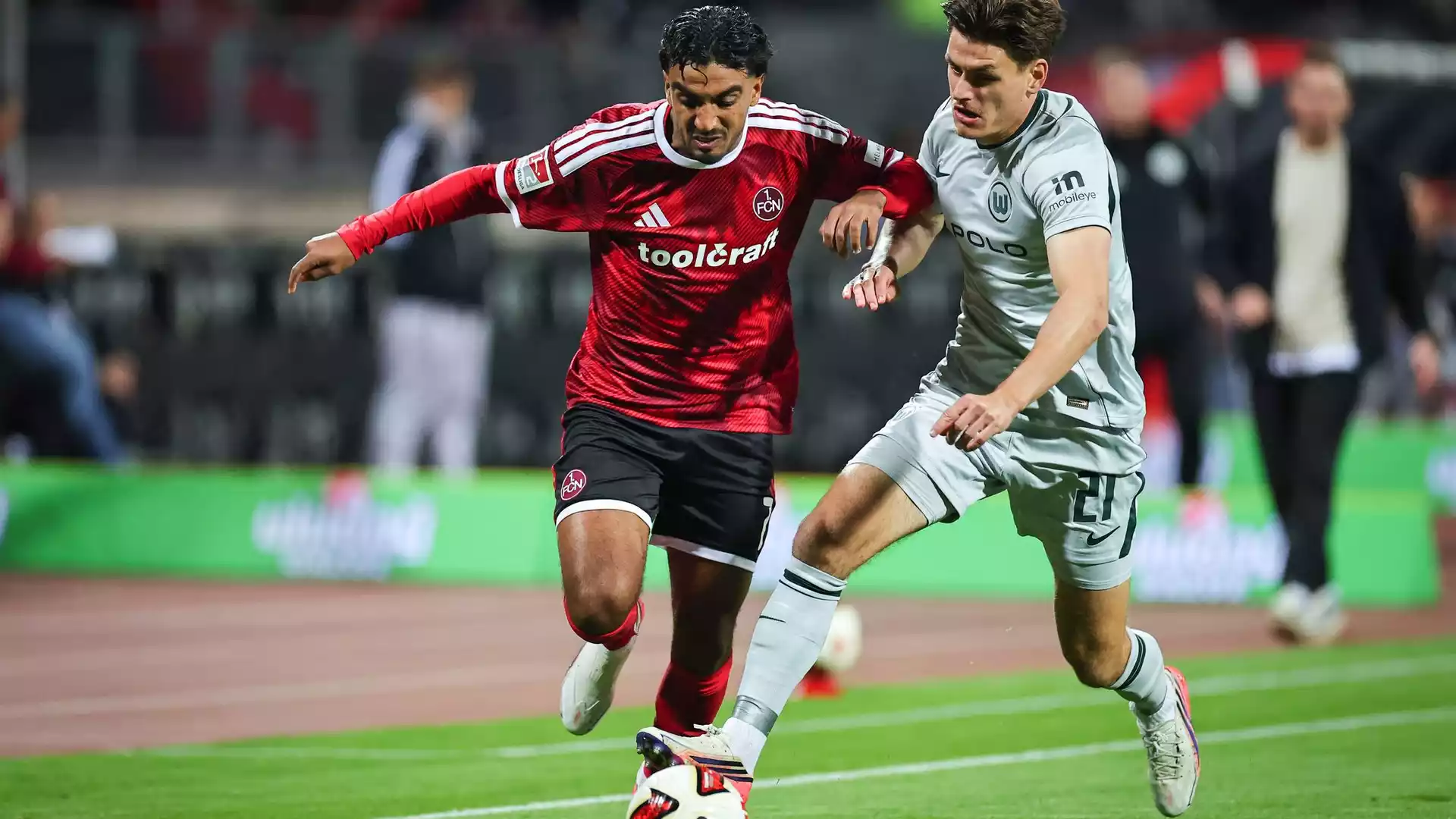 Zwei Fußballspieler von 1. FC Nürnberg und VfL Wolfsburg kämpfen am Ball auf dem Spielfeld.