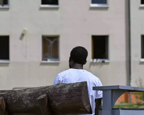 Ein Mann sitzt auf einer Bank vor einem Gebäude und blickt auf die Fassade.