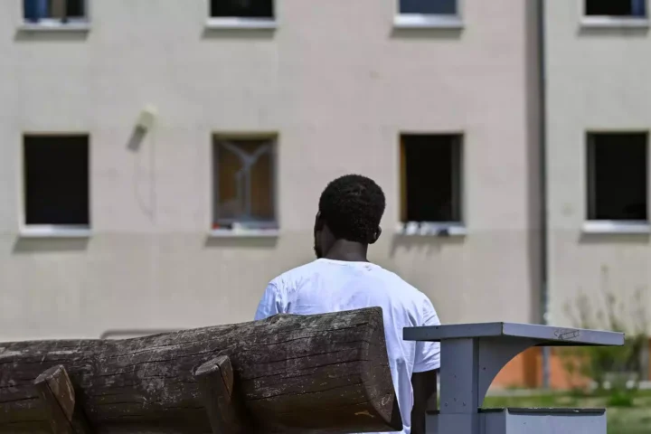 Ein Mann sitzt auf einer Bank vor einem Gebäude und blickt auf die Fassade.