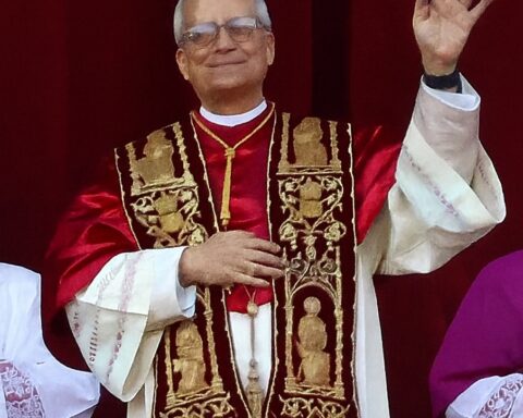 Papst Leo XIV. winkt in liturgischer Kleidung vor rotem Hintergrund.