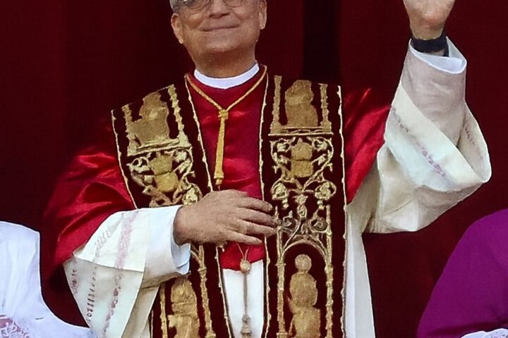 Papst Leo XIV. winkt in liturgischer Kleidung vor rotem Hintergrund.