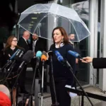 Eine Politikerin spricht vor einem Eingang unter einem transparenten Schirm in ein Mikrofonbündel, umgeben von Journalisten.