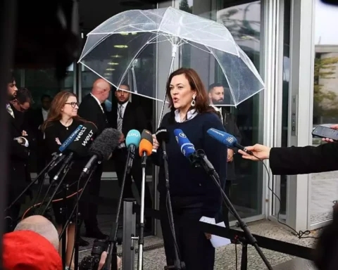 Eine Politikerin spricht vor einem Eingang unter einem transparenten Schirm in ein Mikrofonbündel, umgeben von Journalisten.