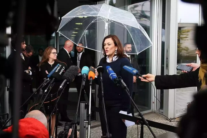Eine Politikerin spricht vor einem Eingang unter einem transparenten Schirm in ein Mikrofonbündel, umgeben von Journalisten.