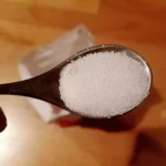 Nahaufnahme eines Löffels mit weißem Zucker in einer Hand.