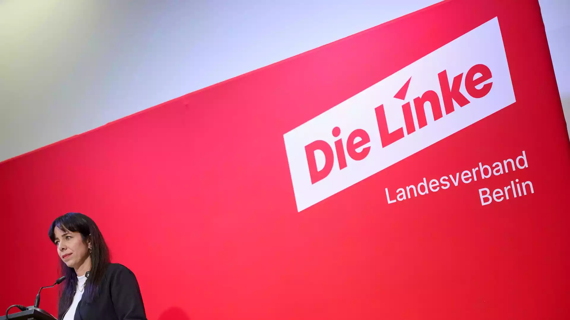 Eine Frau spricht an einem Podium vor einer roten Wand mit dem Logo der Berliner Linken.
