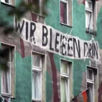 Wohnhausfassade mit großem Protestschriftzug „Wir bleiben drin“.