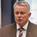 Mann im Anzug spricht in Innenraum, porträtiert vor unscharfem Hintergrund.