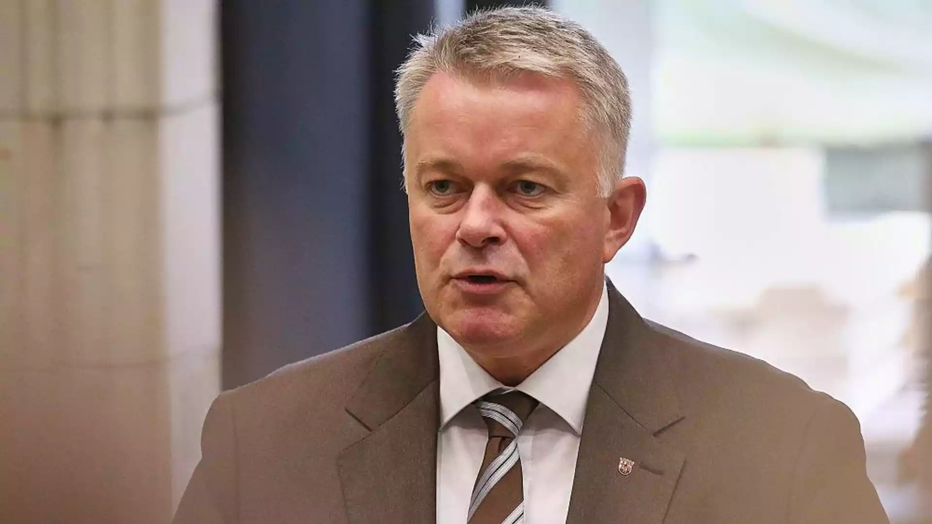 Mann im Anzug spricht in Innenraum, porträtiert vor unscharfem Hintergrund.