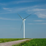Windkraftanlage neben einer Landstraße über grünen Feldern unter blauem Himmel.