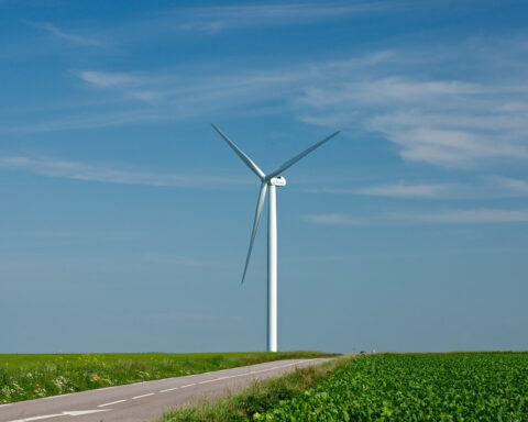 Windkraftanlage neben einer Landstraße über grünen Feldern unter blauem Himmel.