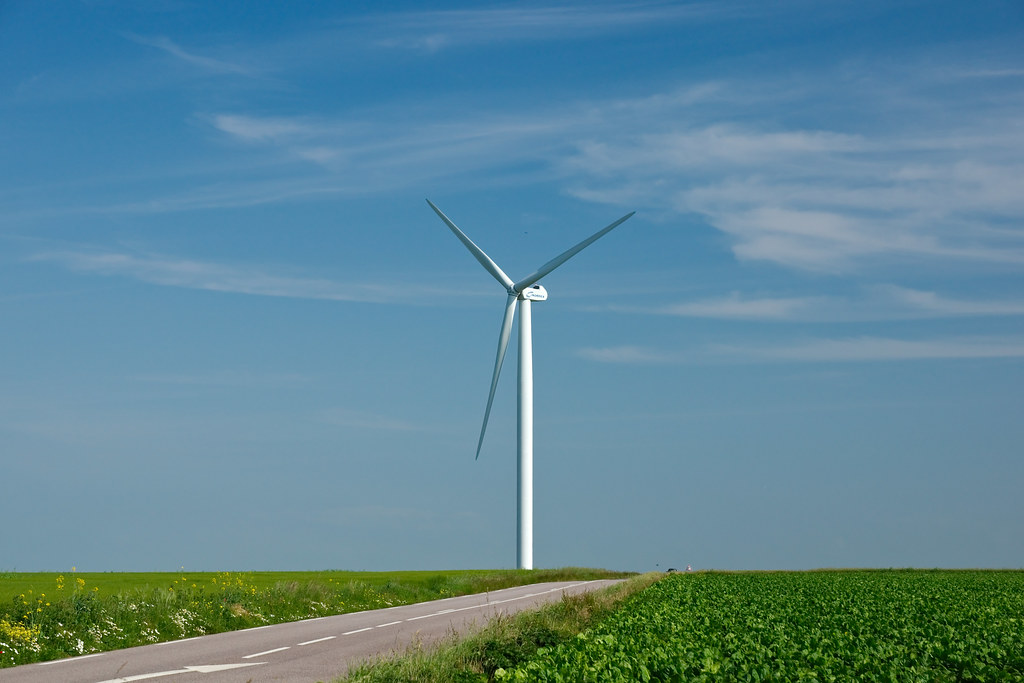 Windkraftanlage neben einer Landstraße über grünen Feldern unter blauem Himmel.