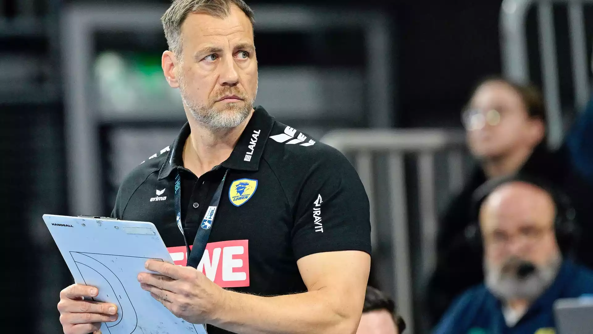 Maik Machulla steht mit Klemmbrett am Spielfeldrand in schwarzem Rhein-Neckar-Löwen-Poloshirt.