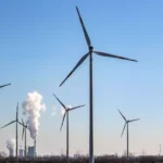 Windräder stehen vor Industrieanlagen mit aufsteigendem Dampf unter blauem Himmel.