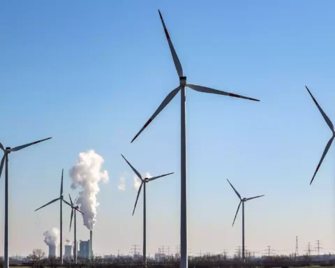 Windräder stehen vor Industrieanlagen mit aufsteigendem Dampf unter blauem Himmel.
