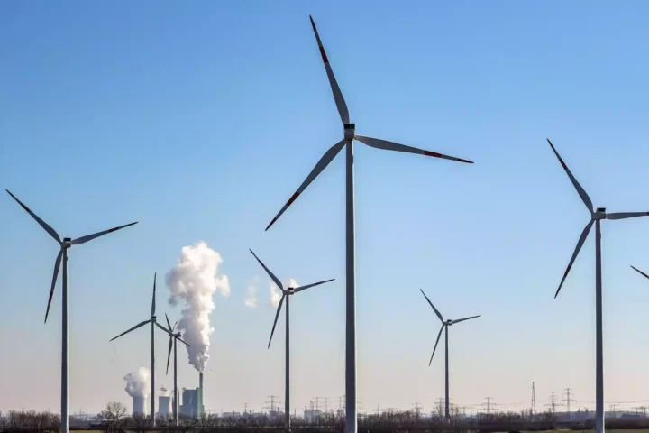 Windräder stehen vor Industrieanlagen mit aufsteigendem Dampf unter blauem Himmel.