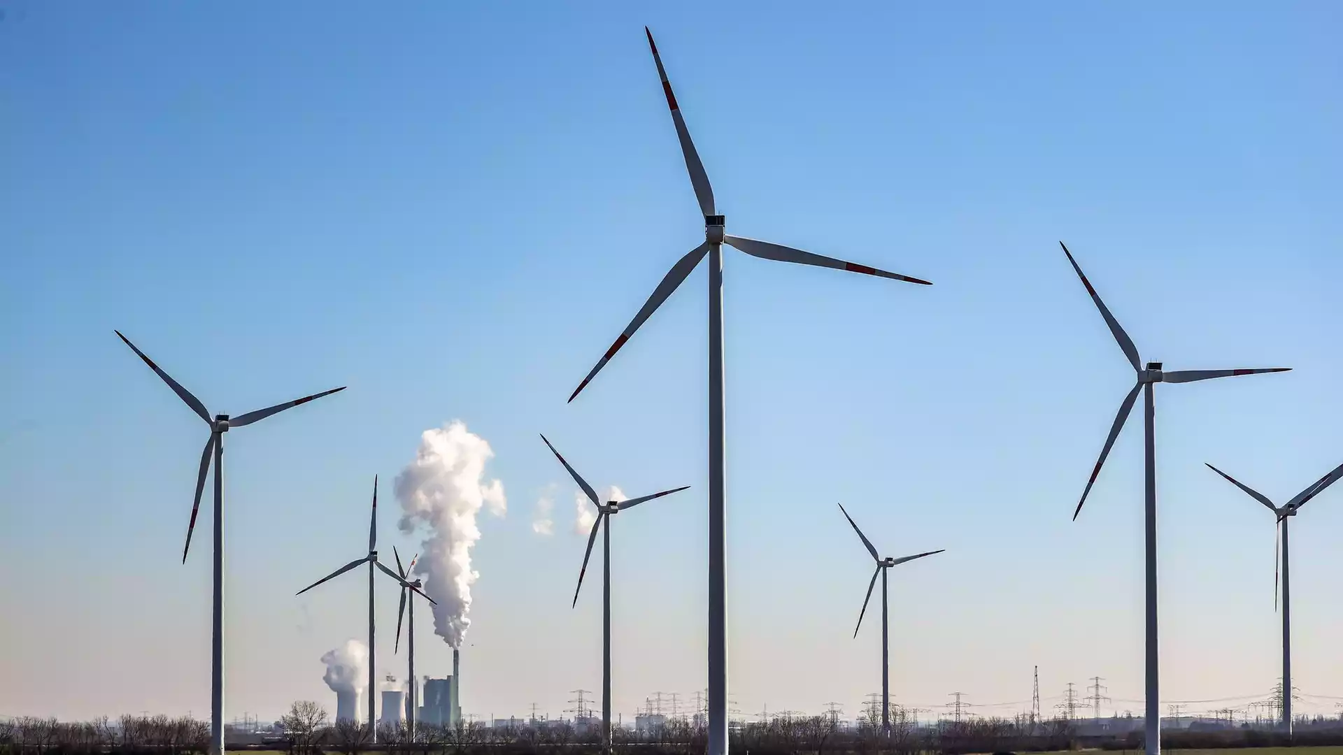 Prognos beziffert Klimaneutralität bis 2045 auf sechs Billionen Euro 1 Windräder stehen vor Industrieanlagen mit aufsteigendem Dampf unter blauem Himmel.