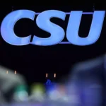 CSU-Logo vor dunklem Hintergrund, unscharfe Personen im Vordergrund.