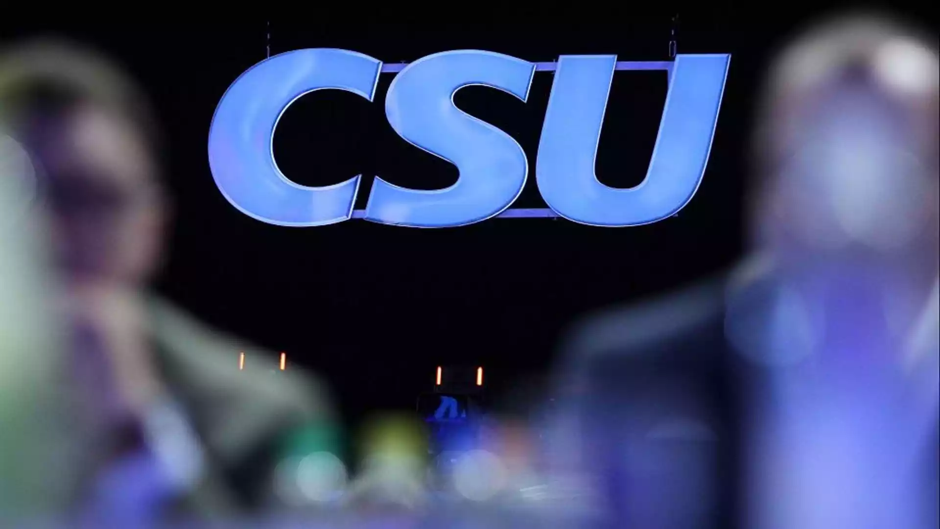 CSU-Logo vor dunklem Hintergrund, unscharfe Personen im Vordergrund.