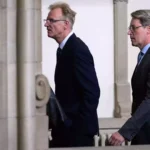 Andreas Scheuer geht in dunklem Anzug durch einen Innenraum mit Säulen.