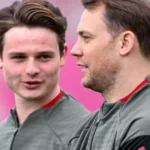 Zwei Fußballspieler im Training in grauen Oberteilen vor pinkem Hintergrund, einer im Profil, der andere blickt nach vorn.