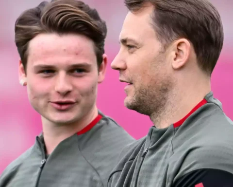 Zwei Fußballspieler im Training in grauen Oberteilen vor pinkem Hintergrund, einer im Profil, der andere blickt nach vorn.