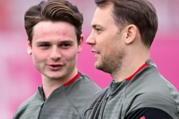 Zwei Fußballspieler im Training in grauen Oberteilen vor pinkem Hintergrund, einer im Profil, der andere blickt nach vorn.