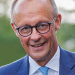Friedrich Merz lächelt in blauem Anzug und mit Brille in einer Porträtaufnahme.