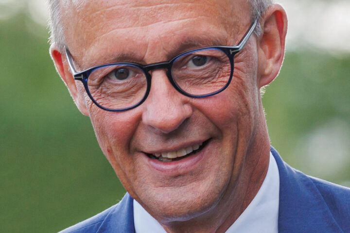 Friedrich Merz lächelt in blauem Anzug und mit Brille in einer Porträtaufnahme.