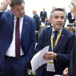 Zwei Männer in Anzügen sprechen in einem Plenarsaal, einer hält Papiere in der Hand.