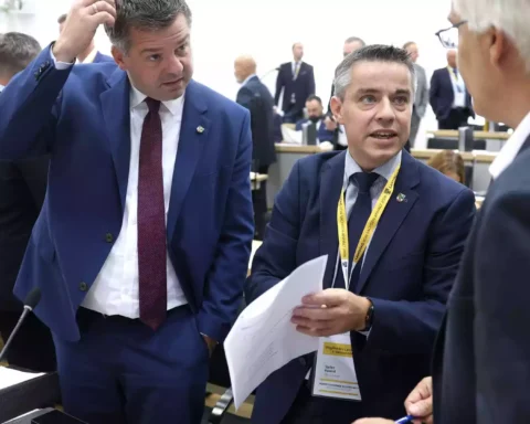 Zwei Männer in Anzügen sprechen in einem Plenarsaal, einer hält Papiere in der Hand.