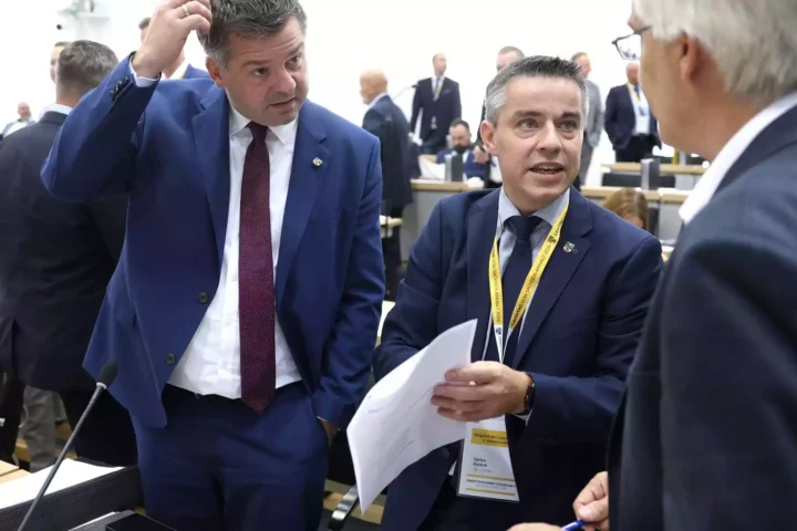 Zwei Männer in Anzügen sprechen in einem Plenarsaal, einer hält Papiere in der Hand.