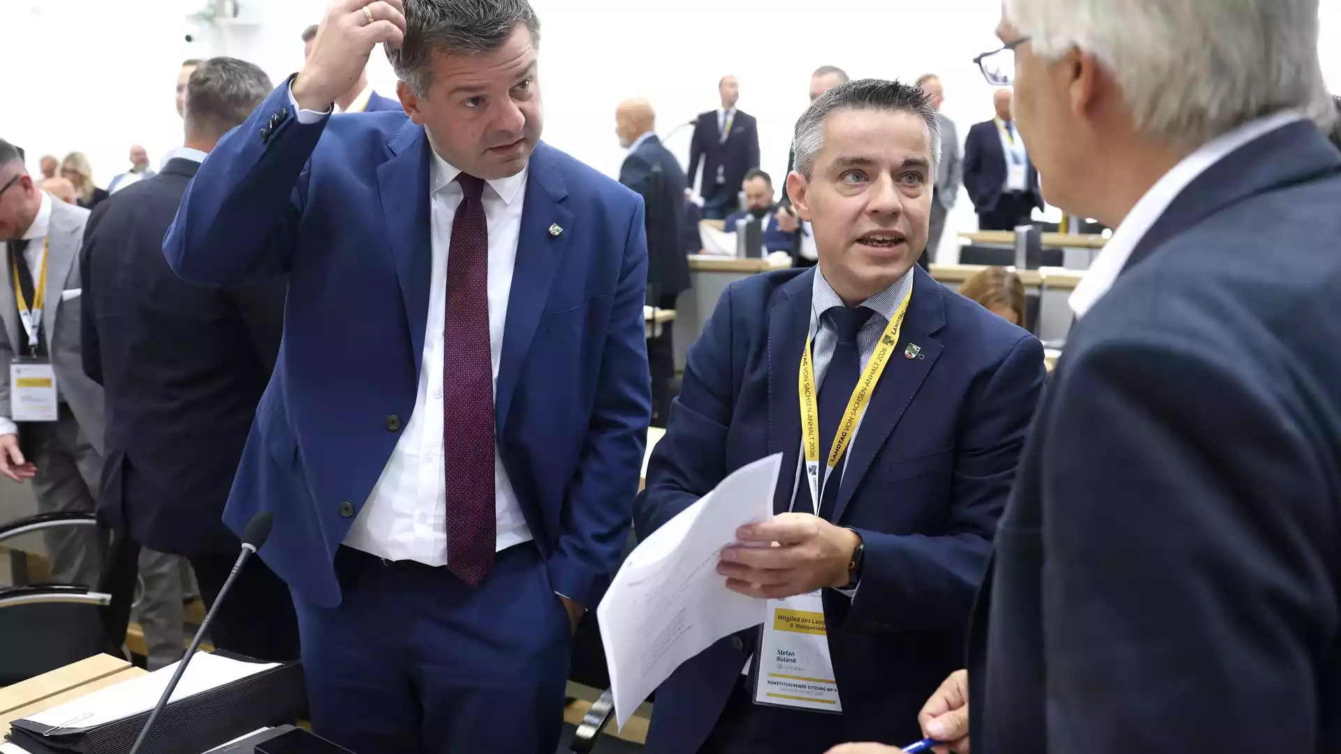 Zwei Männer in Anzügen sprechen in einem Plenarsaal, einer hält Papiere in der Hand.