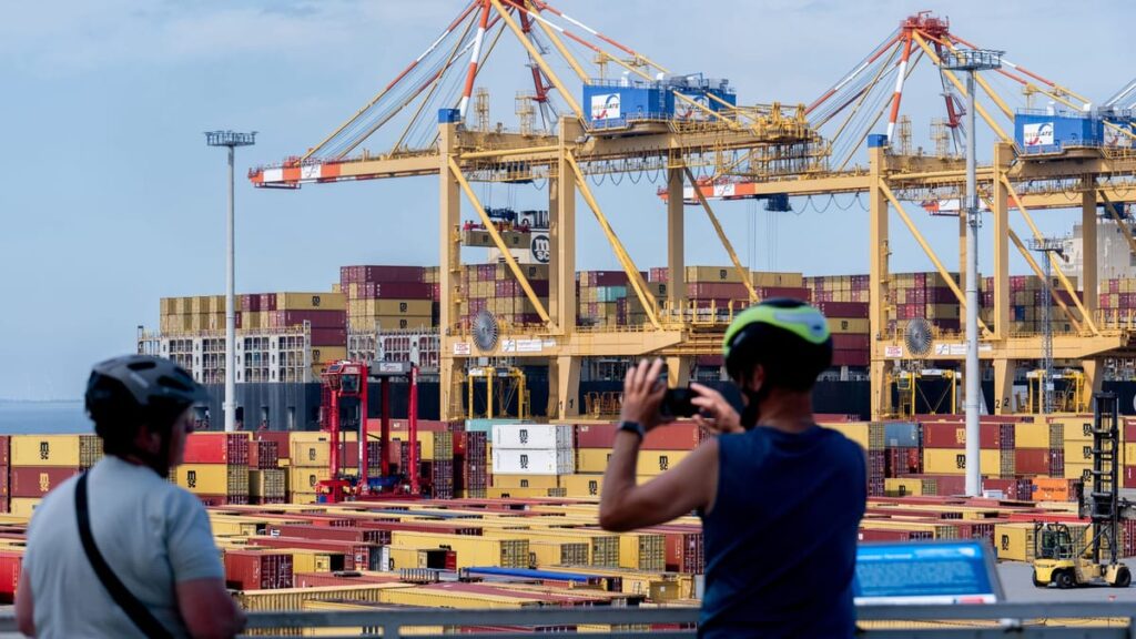Containerhafen mit großen Hafenkranen und gestapelten Containern, im Vordergrund zwei Radfahrer.