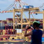 Containerhafen mit großen Hafenkranen und gestapelten Containern, im Vordergrund zwei Radfahrer.