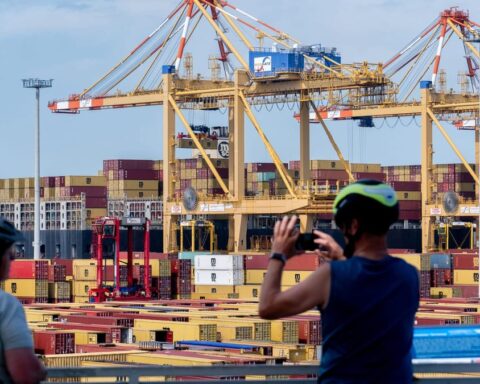Containerhafen mit großen Hafenkranen und gestapelten Containern, im Vordergrund zwei Radfahrer.