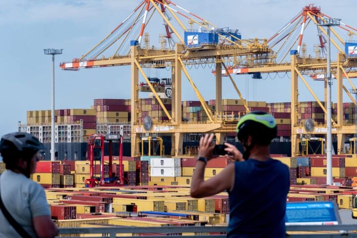 Containerhafen mit großen Hafenkranen und gestapelten Containern, im Vordergrund zwei Radfahrer.